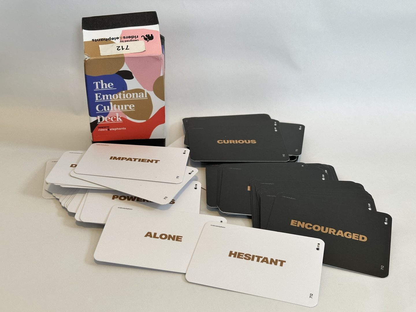 The Emotional Culture Deck box beside spread cards, white and black, each printed with one emotion word such as curious, impatient, encouraged and hesitant.